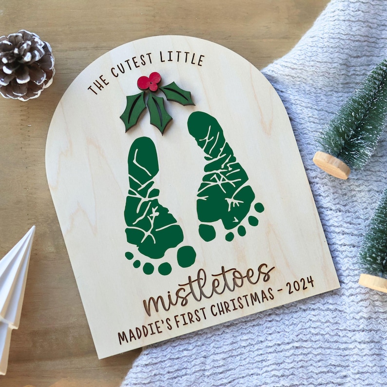 K&ouml;nnte beinhalten: Ein Holzschild mit dem Text "The Cutest Little Mistletoes Maddie's First Christmas - 2024". Das Schild zeigt zwei gr&uuml;ne Fu&szlig;abdr&uuml;cke und ein gr&uuml;nes Blatt mit einer roten Beere.
