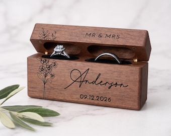 May include: A rectangular dark wood ring box with a hinged lid, featuring laser-engraved floral line art, the text MR & MRS, the name Anderson, and the date 09.12.2026. The box contains two black velvet ring slots holding a diamond ring and a band.