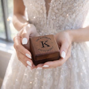 May include: A small, square wooden ring box held in open hands. The box is dark brown with the text "THE KNOWLES" above a large letter "K" and the date "03/26/2021". The box is closed and appears to be new.