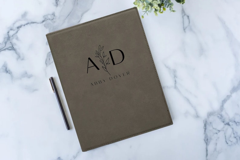 May include: A brown leatherette folder with a floral design and the initials "AD" in black. The name "Abby Dover" is printed below the initials.