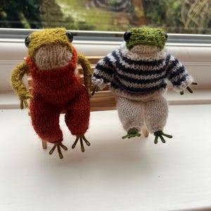 Könnte beinhalten: Zwei handgestrickte Froschfiguren. Einer trägt eine orangefarbene Latzhose und eine gelb-grüne Kopfbedeckung. Der andere Frosch trägt einen blau-weiß gestreiften Pullover und eine weiße Hose. Beide haben schwarze Augen und grüne Füße und sitzen auf einer kleinen Holzbank.