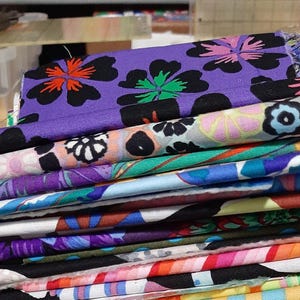 May include: A stack of colorful fabric pieces with floral patterns, including purple, orange, and green designs. The fabric rests on a scale, displaying a weight of 16.7 oz.