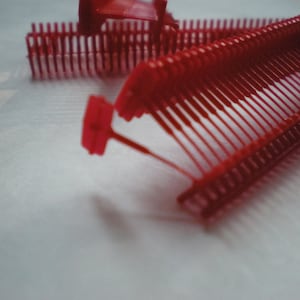 May include: Close-up of several red plastic tag fasteners, also known as tag guns or barbs, used for attaching price tags or labels to clothing and other merchandise. The fasteners are arranged on a white surface.