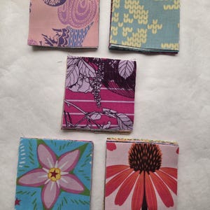 May include: Five stacks of square fabric swatches featuring various patterns. The top swatches display a pink floral print, a light blue knit-style pattern, a dark magenta botanical design, a blue flower with a star center, and a large orange coneflower.