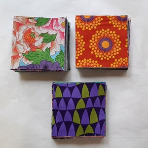 May include: Three stacks of fabric squares with different patterns. One stack features large floral blooms in pink, orange, and green. Another has a red background with yellow and orange circular designs. The third stack has a purple and green geometric pattern.