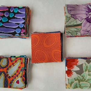 Könnte beinhalten: Stapel bunter Stoffquadrate in verschiedenen Mustern. Die Stoffe zeigen Designs in Lila, Türkis, Orange und Blumenmustern. Die Quadrate sind auf einer weißen Oberfläche angeordnet und zeigen die Vielfalt der Textilien.