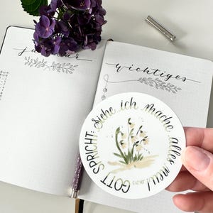 Op de afbeelding: Een open notitieboek met handgeschreven tekst en bloemenillustraties, naast een paarse hortensia en een ronde sticker. De sticker heeft een aquarel illustratie van witte bloemen en Duitse tekst. Een pen en een metalen penhouder zijn ook zichtbaar.