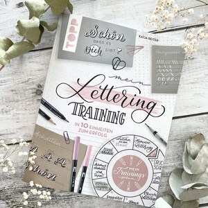 Könnte beinhalten: Ein Buch mit dem Titel "Mein Lettering Training" mit einem rosa-weißen Kreisdiagramm, das verschiedene Lettering-Stile zeigt. Der Buchumschlag hat den Text "In 10 Einheiten zum Erfolg" und "Schatten" darauf geschrieben.