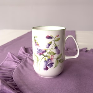 Puede incluir: Taza de cerámica blanca con un diseño floral de flores moradas y rosas y hojas verdes. La taza tiene un borde verde y un asa curva. La taza está sobre una tela morada.