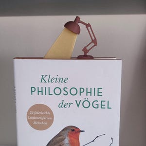 May include: A brown metal bookmark shaped like a desk lamp, with a yellow light beam, is placed on top of a book. The book cover features the title "Kleine Philosophie der Vögel" and an illustration of a bird.