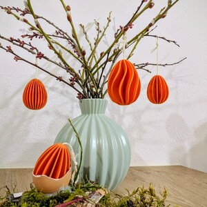 Könnte beinhalten: Eine Osterdekoration mit orangefarbenen, geschichteten Eierornamenten, die an Zweigen in einer hellblauen Vase hängen. Eine Eierschale mit einem orangefarbenen Ei liegt auf Moos und Holz.