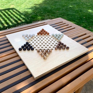 Puede incluir: Un tablero de damas chinas de madera con un patrón de agujeros en forma de estrella. Las piezas del juego son esferas de madera en varios colores: negro, marrón, marrón claro, gris y beige. El tablero está sobre una mesa de madera al aire libre.
