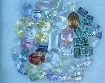 May include: A collection of various gemstones in different shapes and colors, including blue, pink, yellow, and clear. The gemstones are scattered on a light blue surface, showcasing their facets and brilliance. The assortment includes oval, rectangular, and round cuts.