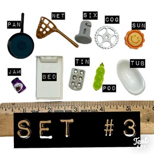 May include: A collection of miniature items labelled PAN, NET, SIX, COG, SUN, JAM, BED, TIN, POD, and TUB, arranged above a wooden ruler showing measurements in inches. The set is identified as SET #3. The items include a black pan, brown net, grey six, silver cog, orange sun, purple jam, white bed, silver tin, green pod, and white tub.