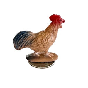 May include: A small plastic rooster figurine with a red comb and wattle, blue and brown feathers, and a brown base. The rooster is standing on a small, round base.