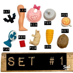 May include: A collection of miniature plastic items including a black bat, a doll leg, a pink bib, a white top, a mug, a blue fan, a red square, a pig, a corn cob, and a walnut. A wooden ruler is visible at the bottom with the text SET # 1.
