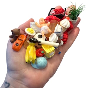 以下が含まれることがあります： テディベア、オレンジ色の車、ポップコーン、赤ちゃん、サッカーボール、バナナ、トマト、アイスクリームコーンなど、ミニチュアのおもちゃのコレクション。 その他のアイテムには、小さな植物、コーヒーカップ、青い野球帽が含まれます。