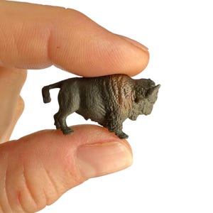 May include: A miniature bison figurine featuring a dark grey body with textured fur detailing and a patch of brown coloration across the shoulder area. The small animal figure is depicted in a standing position with a short tail and small horns.