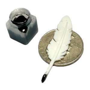 May include: A miniature square inkwell filled with black ink and a small white feather quill resting on a United States quarter coin. The quill tip is dipped in black ink, providing a clear sense of scale for these tiny, detailed objects.