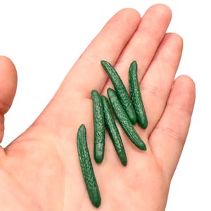 May include: Five small green plastic cucumbers, resembling miniature vegetables, are displayed on an open hand.
