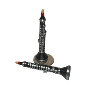 May include: Two miniature black and silver clarinet replicas. One stands upright on a US quarter coin, while the other lies horizontally in front. Both feature detailed silver-colored keys, a black body, and a small red band near the beige mouthpiece.