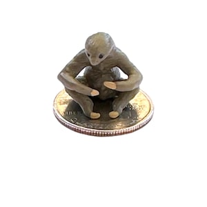 May include: A small, detailed figurine of a seated, humanoid creature with a light brown color. The figurine is resting on a quarter coin, showcasing its small size. The creature has a contemplative pose.