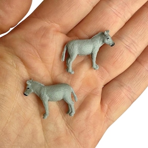 Può includere: Due statuette in miniatura di asino di colore grigio con superfici testurizzate, orecchie a punta e musi scuri. Ogni piccola figura di animale è raffigurata in posizione eretta, mostrando dettagli del pelo modellati e tratti distintivi.