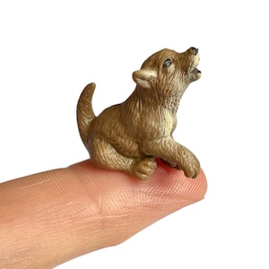 May include: A miniature brown wolf pup figurine with textured fur, depicted in a seated position with its head tilted upward and mouth open as if howling. The small animal figure features pointed ears, a short tail, and detailed paws.