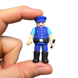 May include: A small police officer figurine wearing a blue uniform with a matching cap, light blue sleeves, a black utility belt, and black boots. The figure has a simple smiling face and is holding a small black baton in one hand.