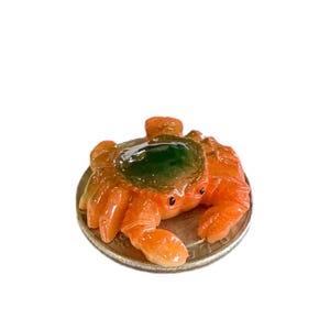 Puede incluir: Una pequeña figurita de cangrejo de cerámica naranja y verde sentada sobre una moneda de 25 centavos.