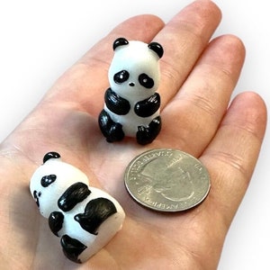 May include: Two small panda figurines, one upright and one lying down, with black and white coloring. A quarter is next to the figurines for size comparison. The pandas have black ears, eyes, and limbs.