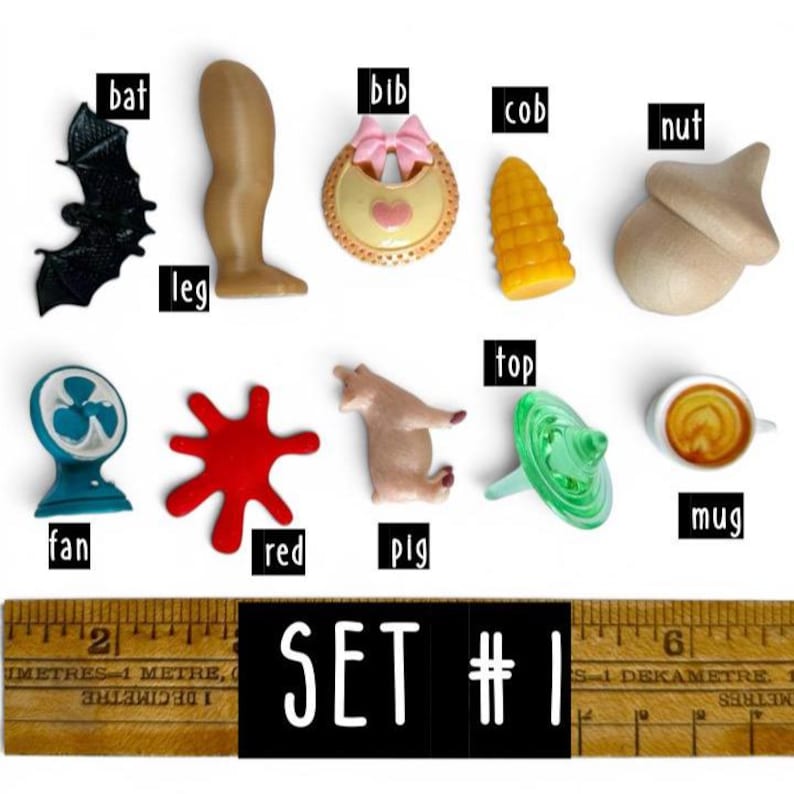 May include: A collection of ten small plastic miniatures labelled bat, leg, bib, cob, nut, fan, red, pig, top, and mug, arranged above a wooden ruler showing a length of approximately 18 cm. The items vary in colour and shape, including a black bat, a tan leg, a yellow bib, a yellow corn cob, a tan nut, a blue fan, a red splash, a tan pig, a green top, and a white mug.