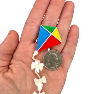 May include: A miniature colorful kite with red, blue, green, and yellow segments, connected to a string of white bows. A U.S. quarter is next to the kite for size comparison. The kite is held in a person's hand.