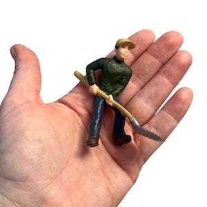 May include: A small plastic figurine of a person wearing a green sweater, blue jeans, and a brown cap, holding a shovel with a wooden handle.