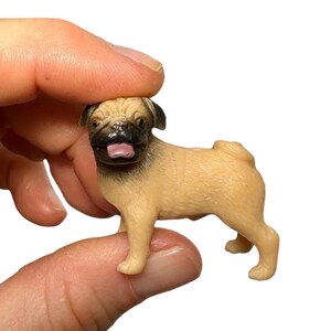 May include: A miniature pug figurine featuring a light tan body with a dark brown muzzle and ears. The small dog figure is depicted in a standing position with its mouth slightly open, revealing a pink tongue, and a curled tail on its back.