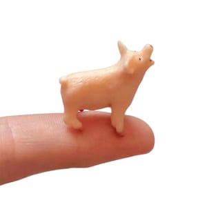 May include: A miniature light pink pig figurine with a slightly open mouth, standing on a finger. The pig has small ears and a short tail. The background is plain white.