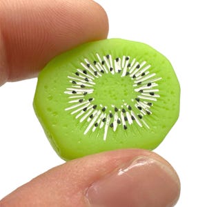 Pode incluir: Uma fatia de kiwi verde com detalhes brancos e pretos. A fatia de kiwi é segurada entre dois dedos contra um fundo branco. O kiwi tem um anel externo verde brilhante e um centro branco com sementes pretas.