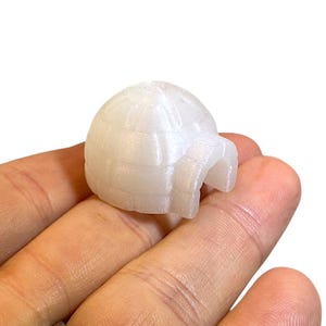May include: A small, white, dome-shaped igloo figurine with a textured block pattern on its exterior surface. The object features a rounded roof and a small, arched entrance opening at the front, crafted from a smooth, slightly pearlescent material.