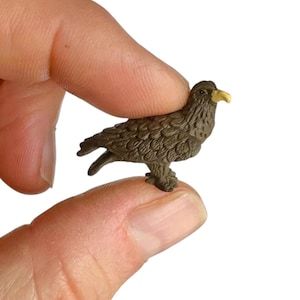 May include: A miniature brown eagle figurine with detailed feather textures and a light yellow beak. The small bird sculpture is depicted in a standing position with its head turned slightly to the side, showcasing a compact and realistic design.