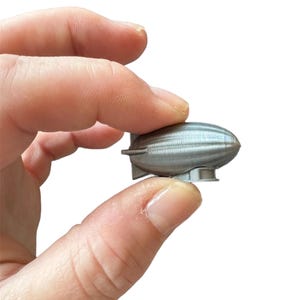 Pode incluir: Um pequeno modelo de dirigível prateado. A pequena aeronave tem um corpo arredondado com linhas horizontais e uma pequena base. O dirigível é segurado entre os dedos contra um fundo branco.
