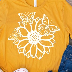 Pode incluir: Uma camiseta amarela mostarda com um gr&aacute;fico branco de um girassol e tr&ecirc;s borboletas.