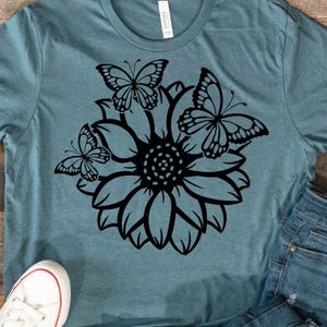 Pode incluir: Uma camiseta azul turquesa com uma silhueta preta de um girassol e tr&ecirc;s borboletas.