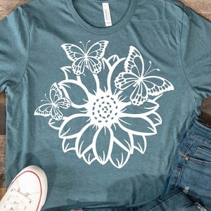 Pode incluir: Uma camiseta azul turquesa com um design gr&aacute;fico branco de um girassol com tr&ecirc;s borboletas ao redor.