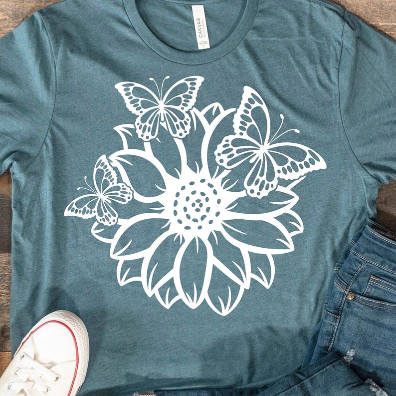Pode incluir: Uma camiseta azul turquesa com um design gr&aacute;fico branco de um girassol com tr&ecirc;s borboletas ao redor.