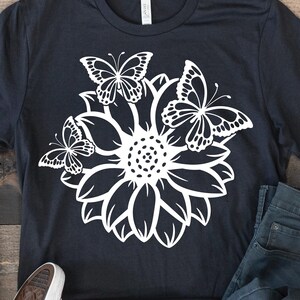 Pode incluir: Uma camiseta preta com um desenho branco de um girassol com tr&ecirc;s borboletas ao redor.