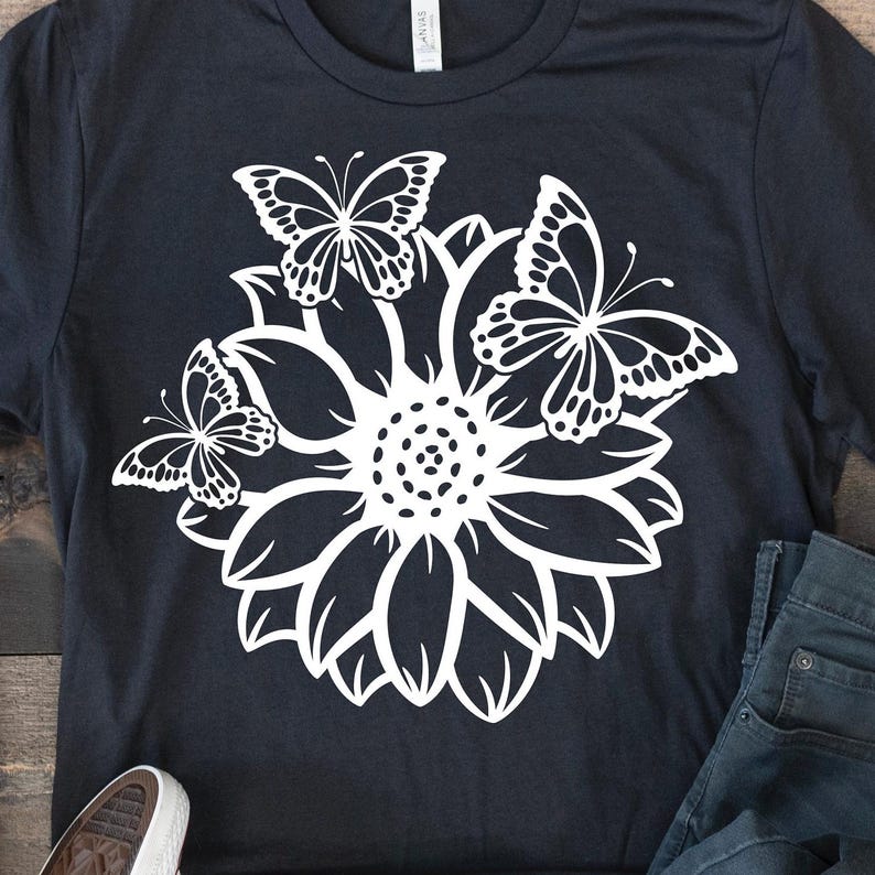 Pode incluir: Uma camiseta preta com um desenho branco de um girassol com tr&ecirc;s borboletas ao redor.