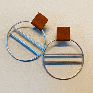 May include: A pair of silver hoop earrings with a brown leather accent. The hoops have a geometric design with two parallel lines.