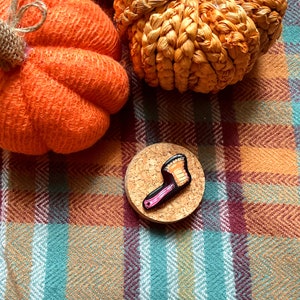 May include: A pink and orange enamel pin shaped like a hand holding a small orange pumpkin. The pin is on a cork background.
