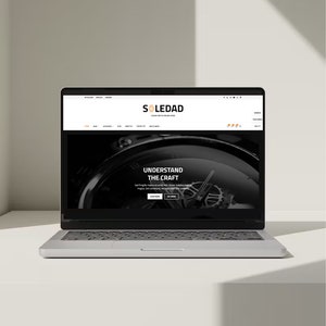 以下が含まれることがあります： 「SOLEDAD」という高級時計店のウェブサイトを表示しているラップトップコンピューター。ウェブサイトには、時計の白黒画像と「UNDERSTAND THE CRAFT」というテキストが表示されています。