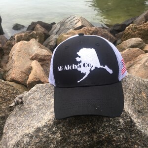 Pode incluir: Boné de caminhoneiro preto e branco com um contorno branco do estado do Alasca e o texto "AK Aloha Co." na frente. O boné tem uma parte de trás de malha branca com uma pequena bandeira americana.